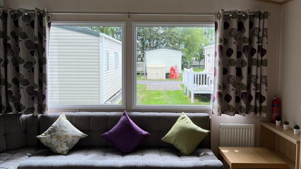 A cozy living room with a grey sofa, three cushions, a wooden table, and a view of a white mobile home through large windows. - Cabin - Sleeps 6 - Beach 10 Minutes Drive (Clacton-on-Sea)