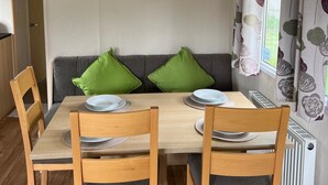 A dining area with a wooden table set for four, two green cushions on a grey sofa, and a potted plant on the wall. - Cabin - Sleeps 6 - Beach 10 Minutes Drive (Clacton-on-Sea)