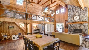 Interior rústico de una cabaña con una mesa de madera, sillas, una chimenea de piedra y una zona superior con una escalera.