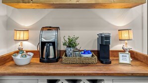Eine Kaffeestation mit zwei Keurig-Kaffeemaschinen, einer Kaffeekanne, einer Schale mit Kaffee-Kapseln, einer Topfpflanze und einem Schild mit dem Wi-Fi-Passwort.