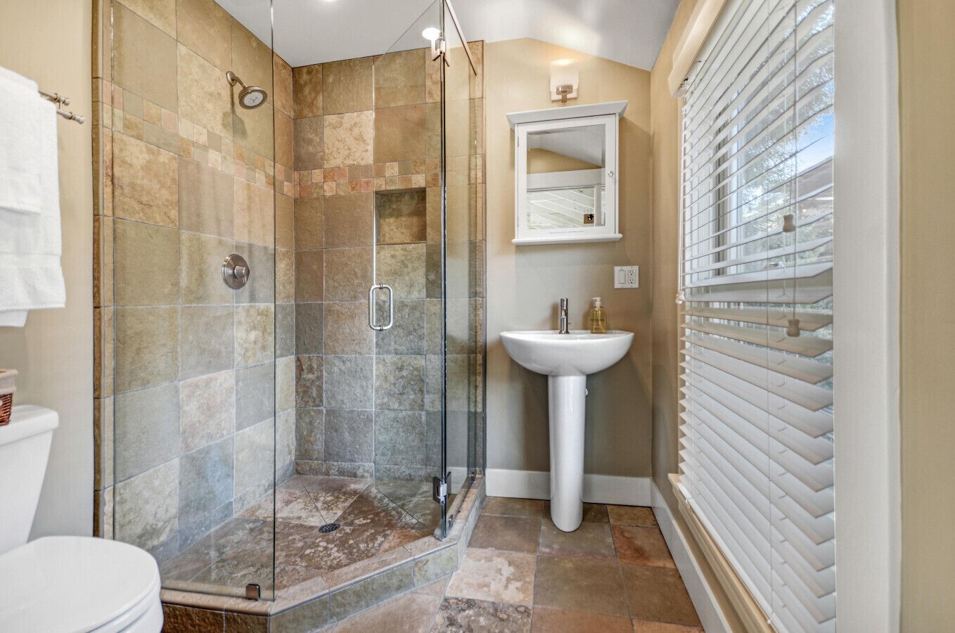 A bathroom with a glass-enclosed shower, a white pedestal sink, and a window with blinds.