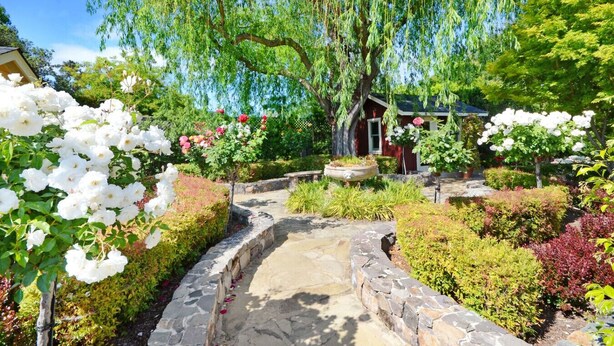 Ein Gartenweg führt zu einem rustikalen Gebäude mit rotem Dach, umgeben von üppiger Bepflanzung und blühenden Blumen.