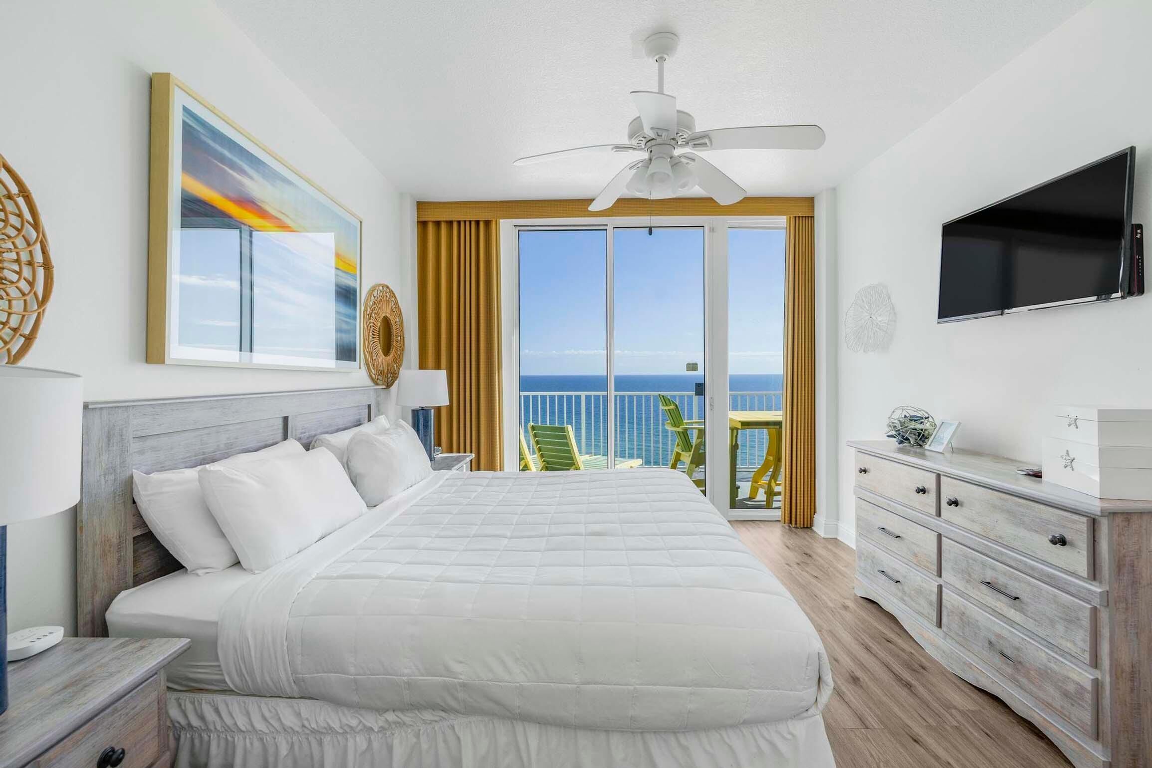 Ein Hotelzimmer mit einem großen Bett, einem Deckenventilator, einem Fernseher, einem Schminktisch und einem Balkon mit Meerblick.