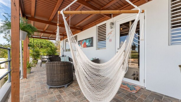 Un hamac suspendu sous un plafond en bois, une chaise en osier et une terrasse carrelée avec une porte et des fenêtres.