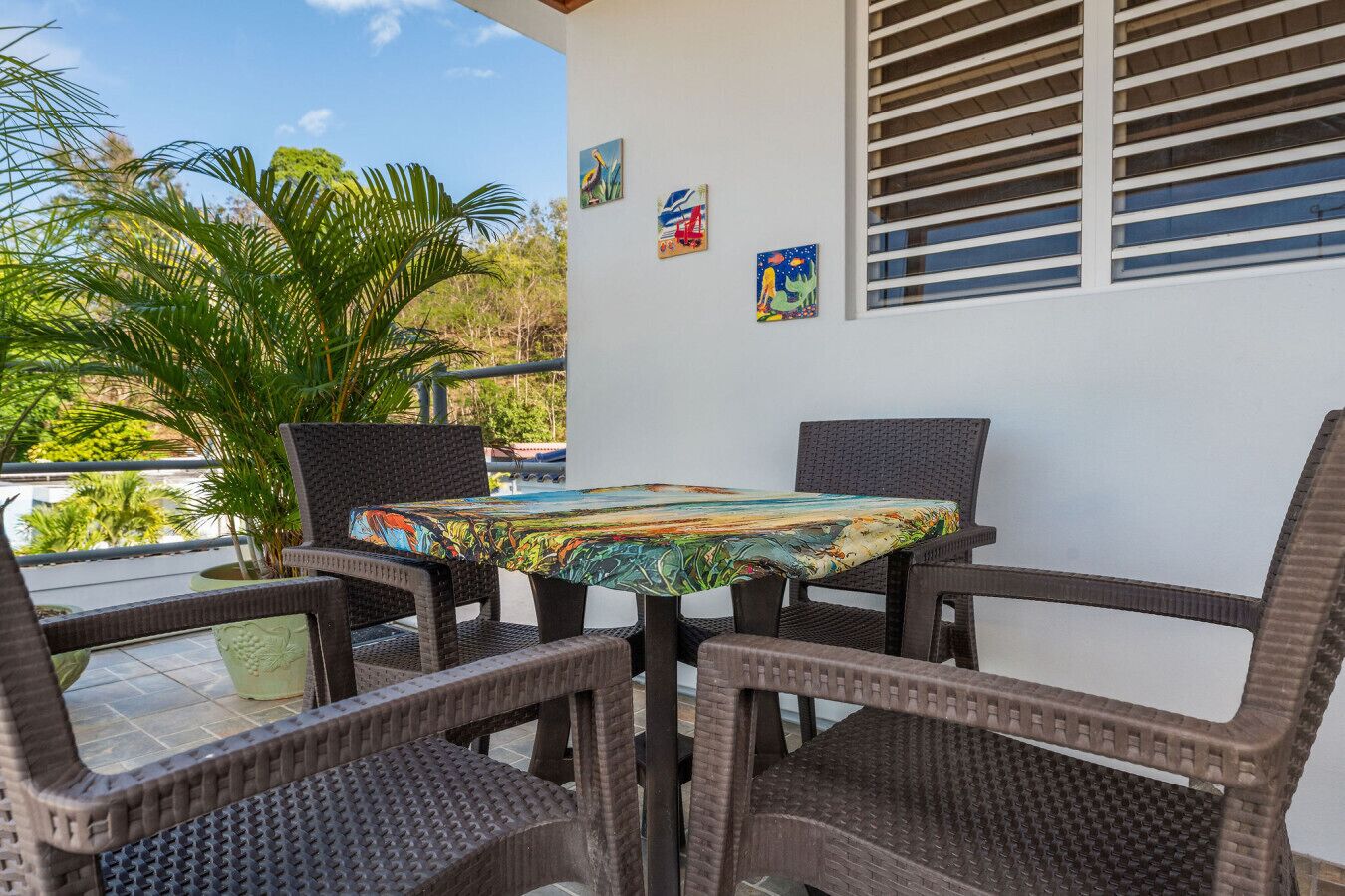 A patio with wicker chairs and a table covered with a colorful tablecloth, surrounded by potted plants and artwork on the wall.