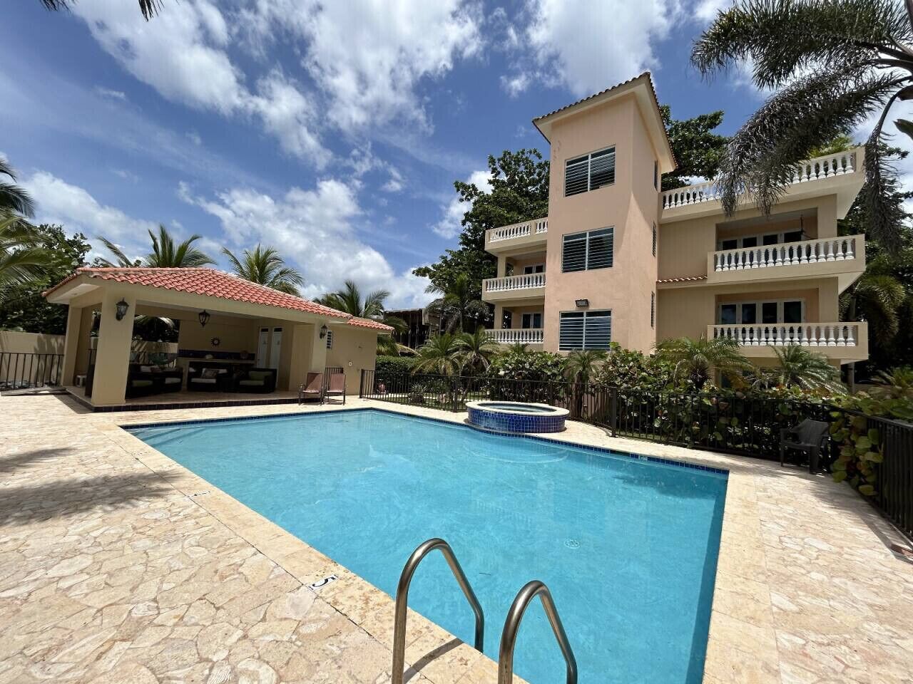 A swimming pool with a stone deck, a covered seating area, and a multi-story building with balconies.