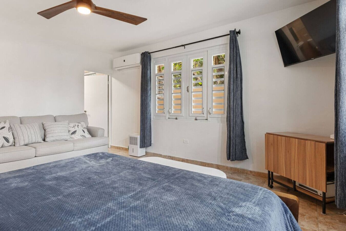Ein Schlafzimmer mit einem Bett, einem Deckenventilator, einem Fernseher, einem Fenster mit Fensterläden und einem Holzkasten.