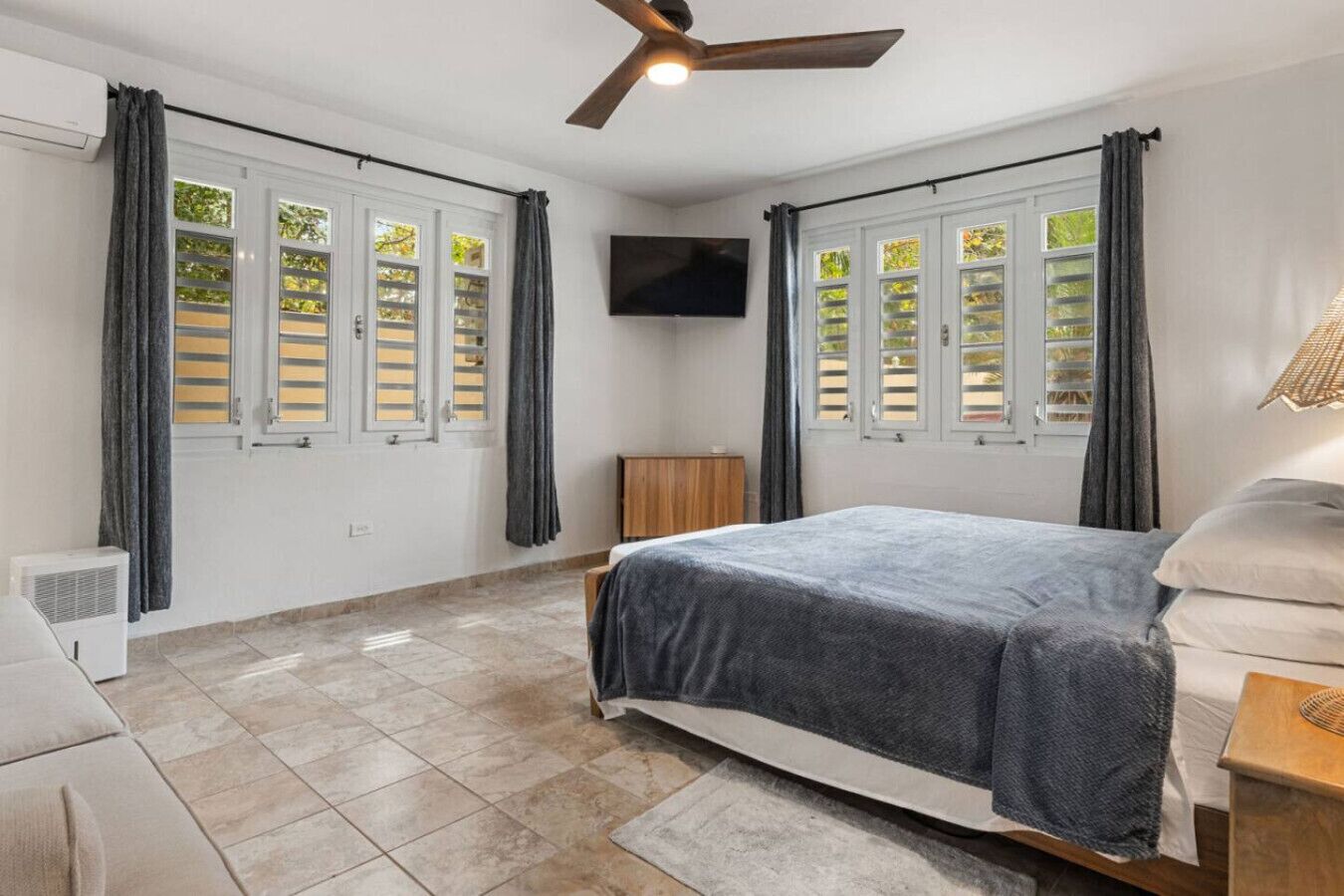 Quarto com cama, ventilador de teto, televisão e janelas com persianas.