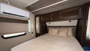 A bedroom with a bed, a window with blinds, a wall-mounted air conditioning unit, and wooden cabinets.