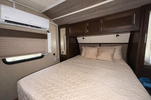 A bedroom with a bed, a window with blinds, a wall-mounted air conditioning unit, and wooden cabinets.