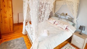 En seng med baldakin og blomstret sengetøj, trægulv, et natbord med lampe og en radiator.