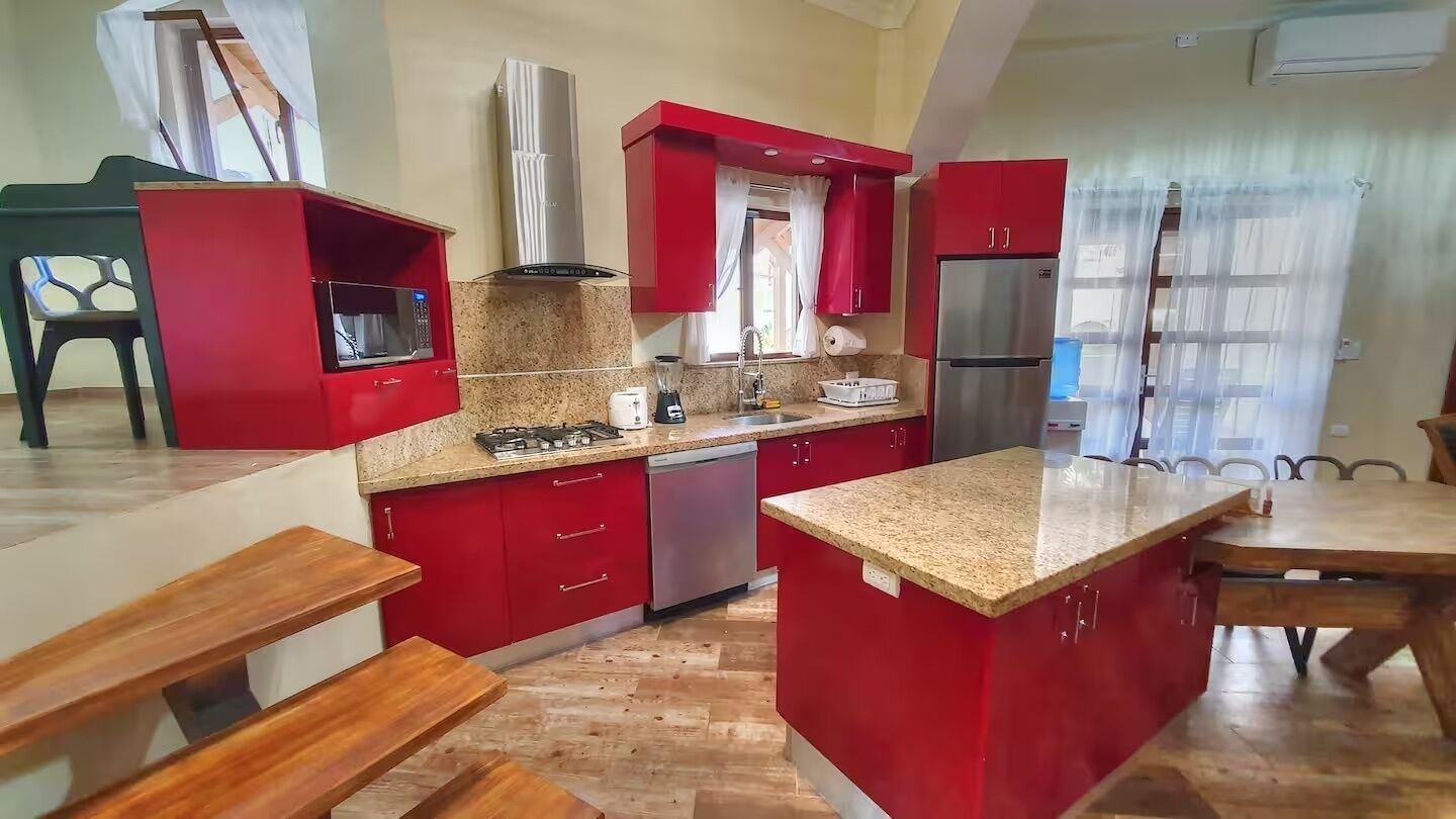 A kitchen with red cabinets, a gas stove, a refrigerator, and a dining area with a wooden table and benches.