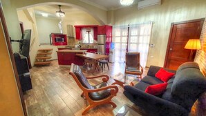 A spacious living area with a wooden floor, a dark blue sofa with red cushions, a wooden coffee table, and a leather armchair. A kitchen with red cabinets and a stainless steel refrigerator is visible in the background. - Cabarete Center 2 BR Condo with a Cozy Terrace (Cabarete)