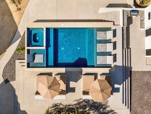 Vue aérienne d’une piscine rectangulaire avec un jacuzzi, entourée de chaises longues et de parasols.