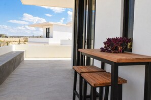 A modern outdoor patio with a wooden table and benches, a potted plant, and a view of a white building and landscape.