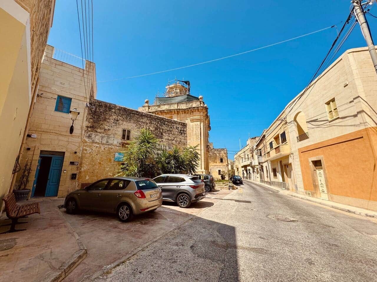 Une rue étroite, bordée de voitures garées, et un bâtiment coiffé d’un dôme en arrière-plan.