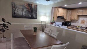 A dining area with a wooden table and white chairs, a painting on the wall, and a kitchen in the background.