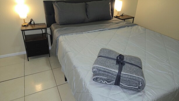 A neatly made bed with a grey blanket and a folded towel, two bedside lamps, a small wooden nightstand, and a dark headboard.