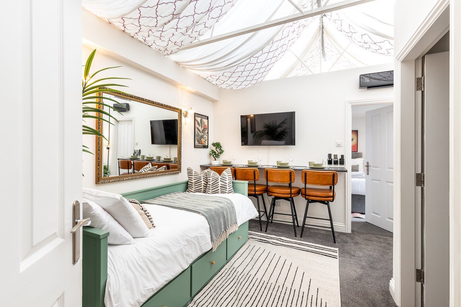 A modern bedroom with a green bed, a large mirror, a dining area with bar stools, and a TV on the wall.