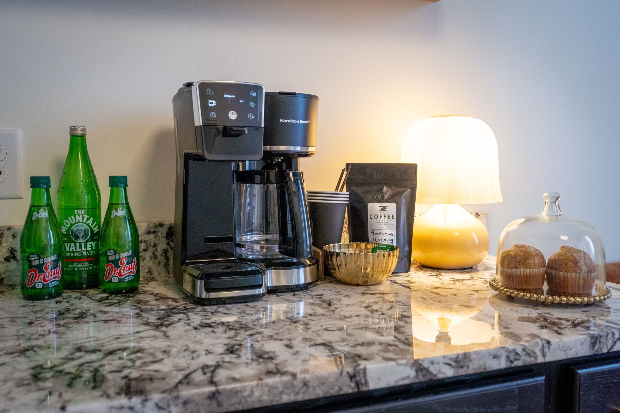 Un comptoir de cuisine avec une machine à café, trois bouteilles d’eau de source Mountain Valley, une bougie et un saladier en verre contenant des muffins.