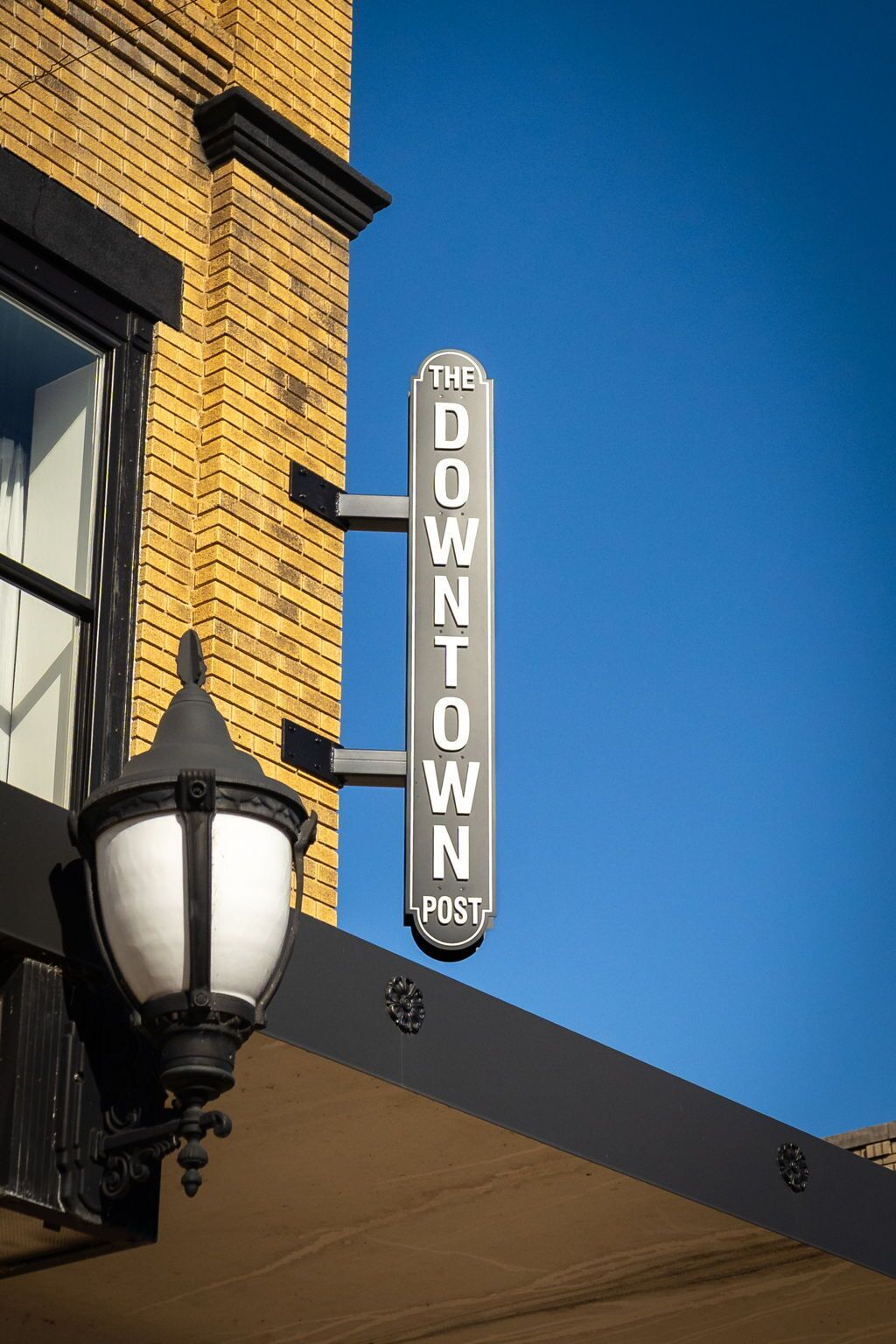 Una señal de calle para 'The Downtown Post' montada en un edificio de fachada de ladrillo y toldo negro.