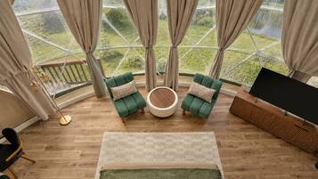 A room with a large window, two green armchairs, a round coffee table, a TV stand, and a wooden floor.