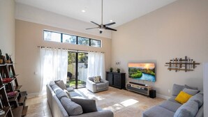 A spacious living room with a large TV, a ceiling fan, and a sliding glass door to a patio. - 3-Bedroom Luxury House (Orlando)