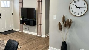A modern living room with a flat-screen TV mounted on a wall, a clock, and a vase with pampas grass. - Modern 3BR Home Near Wheaton College & Cantigny (Wheaton)