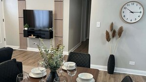 A dining area with a wooden table set for four, surrounded by black chairs. A black vase with greenery is on the table, and a clock is on the wall. - Modern 3BR Home Near Wheaton College & Cantigny (Wheaton)