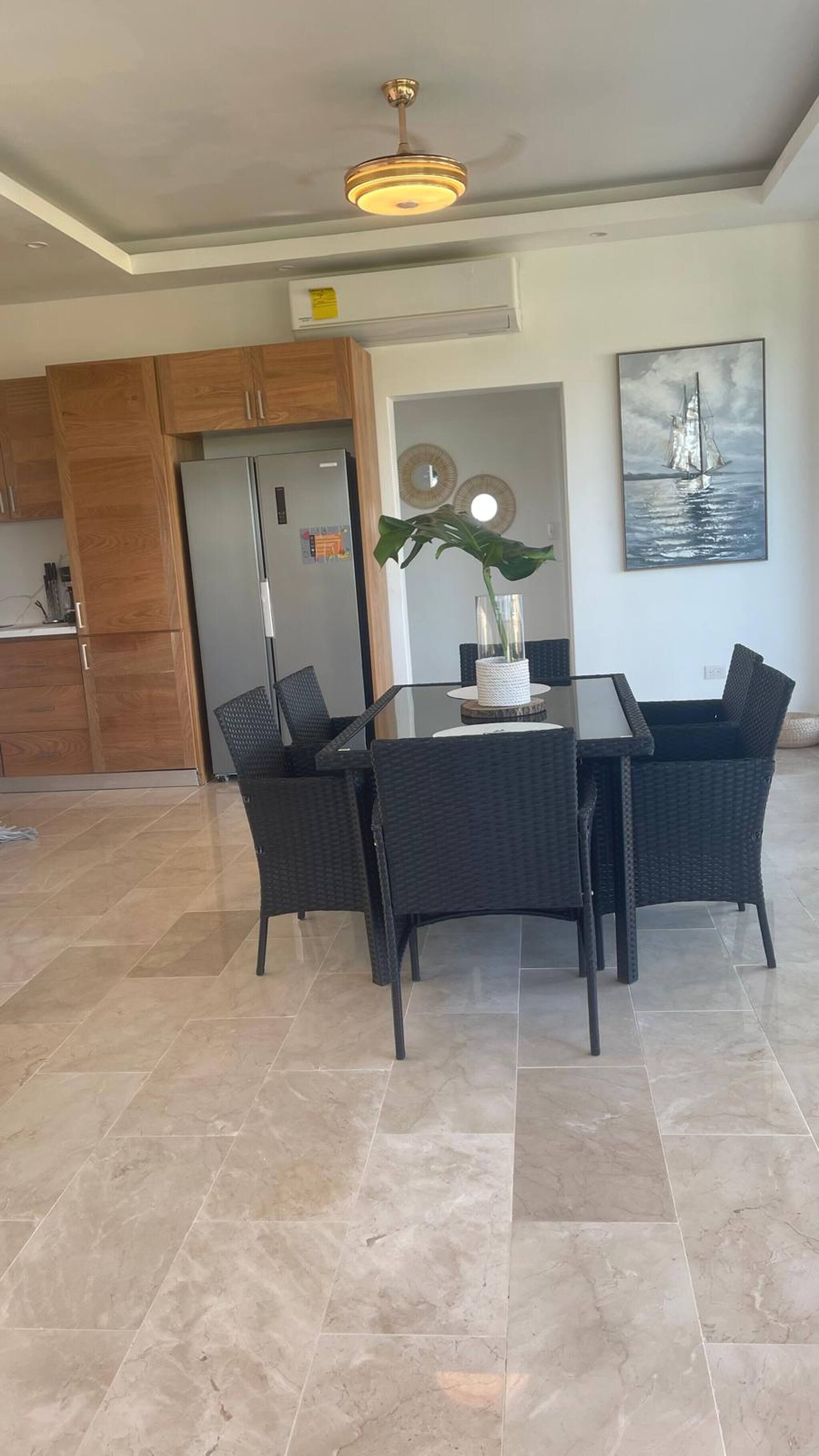 Un coin repas avec une table et des chaises, un ventilateur de plafond et une cuisine en arrière-plan.