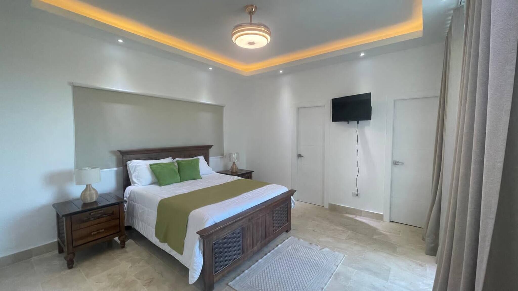 Un dormitorio con cama, mesitas de noche, televisor montado en la pared y una puerta.