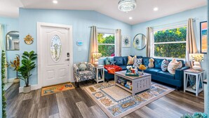 A living room with a blue sofa, a coffee table, and a chandelier. There are windows with curtains, a door with a glass panel, and a rug with a sea theme.