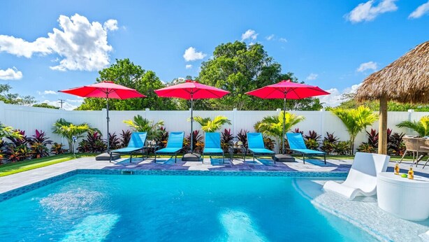 A swimming pool with blue lounge chairs and red umbrellas, surrounded by tropical plants and a white fence.