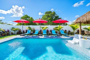A swimming pool with blue lounge chairs and red umbrellas, surrounded by tropical plants and a white fence. - Tiki Hut, Htd Pool, Bar & Putting Greens Near Ave (Delray Beach)