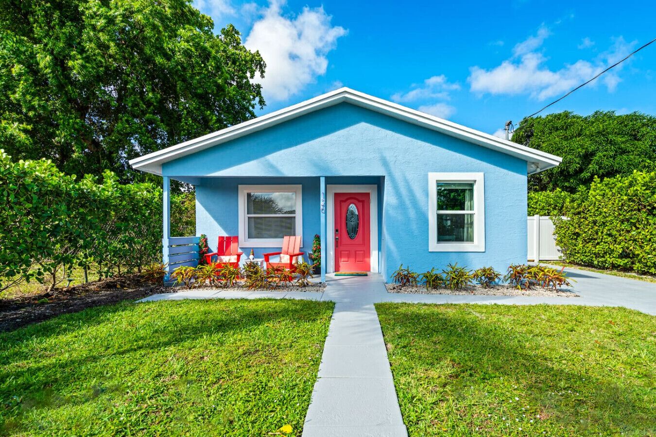 A small, single-story house with a blue exterior, a red door, and a front porch with red chairs.
