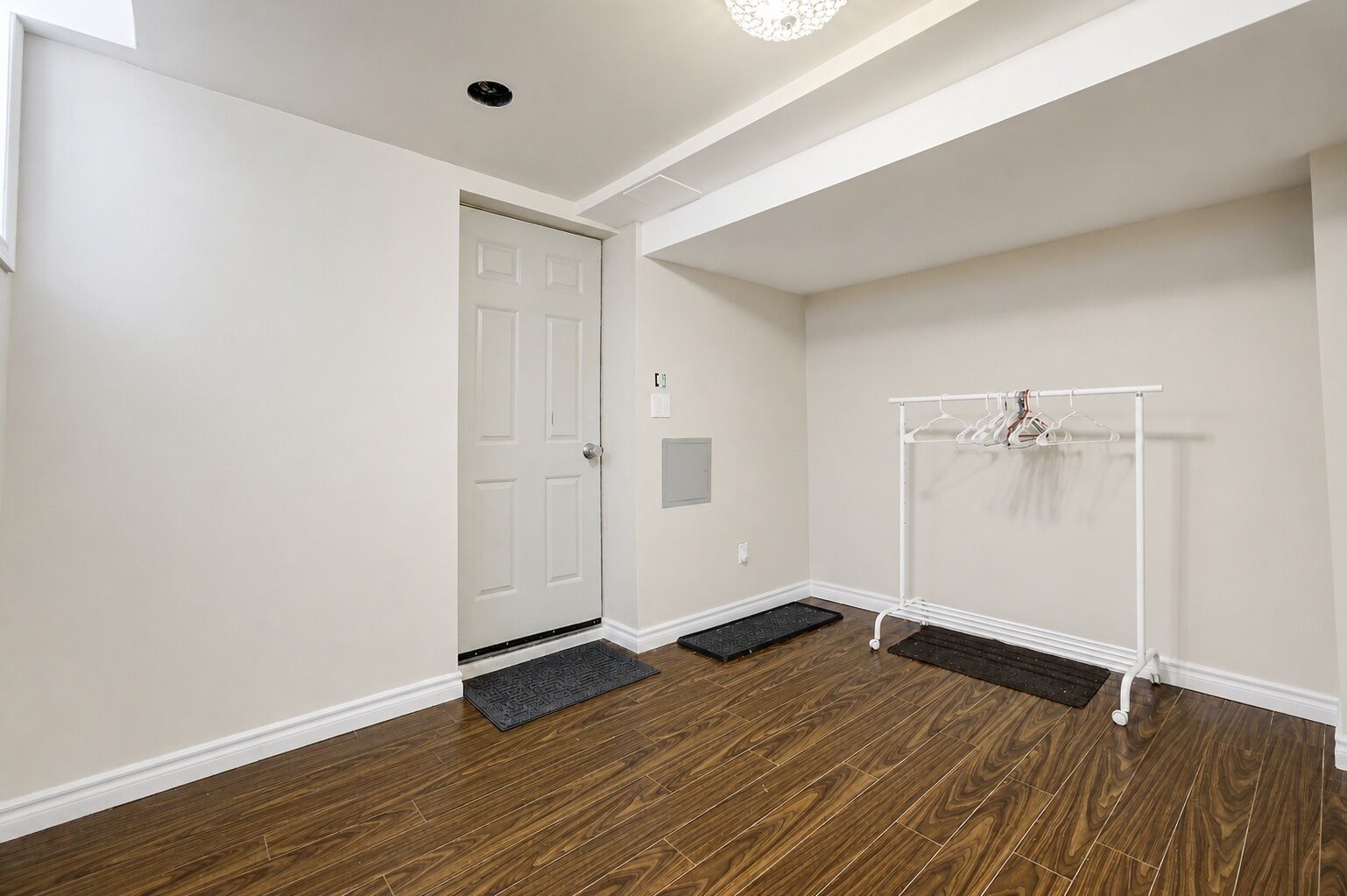 Une pièce avec un plancher en bois, une porte blanche, un porte-manteau avec des cintres et un petit paillasson à l’entrée.