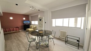 Uma sala de estar moderna com uma mesa de vidro, duas cadeiras, um sofá, uma TV e uma janela com persianas.