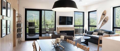 A modern interior with a dining area, sofas, a fireplace, and large windows offering a view of the outdoors.