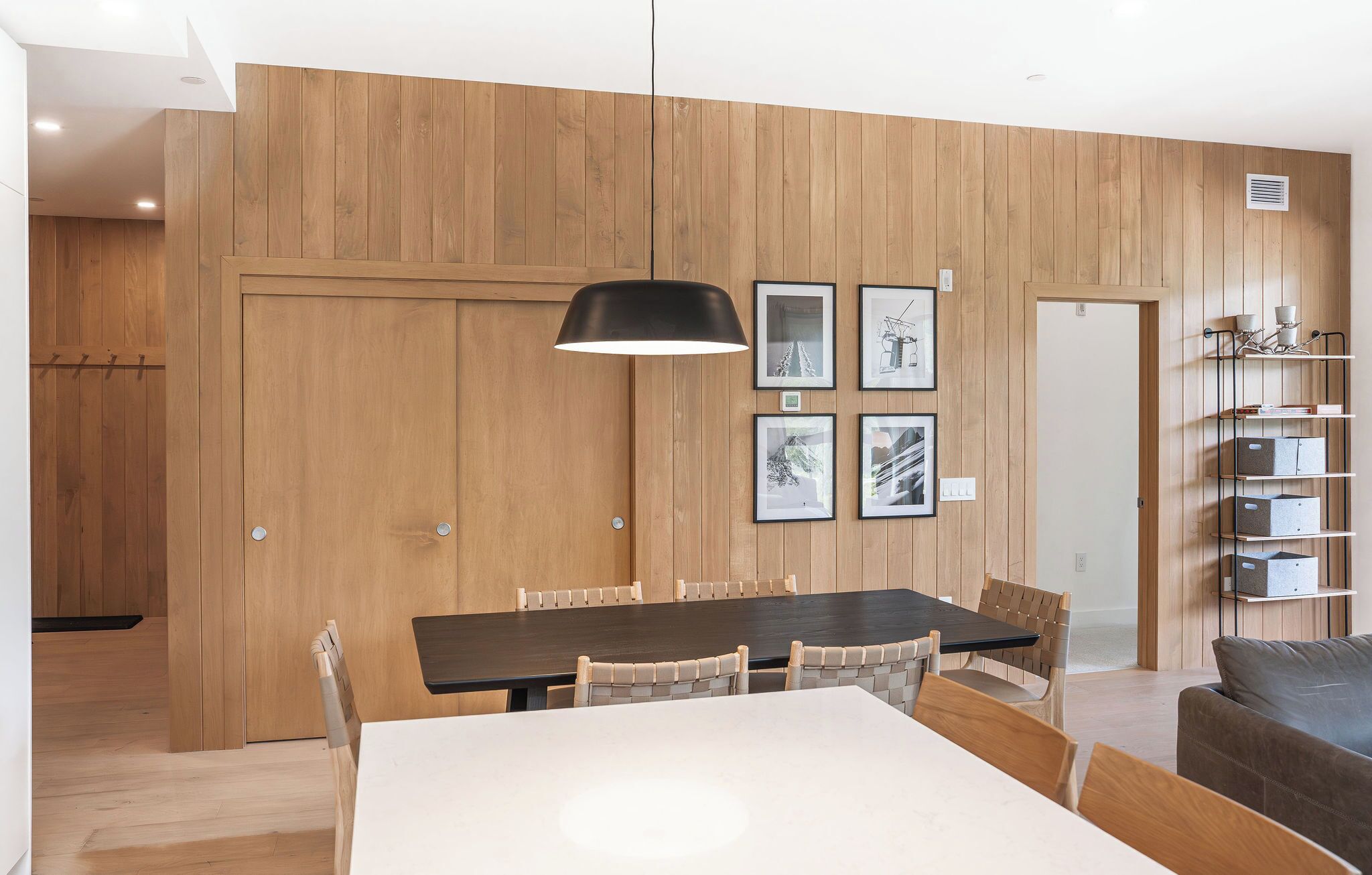 A modern dining area with a wooden paneled wall, a dining table with chairs, a pendant light, and framed pictures.