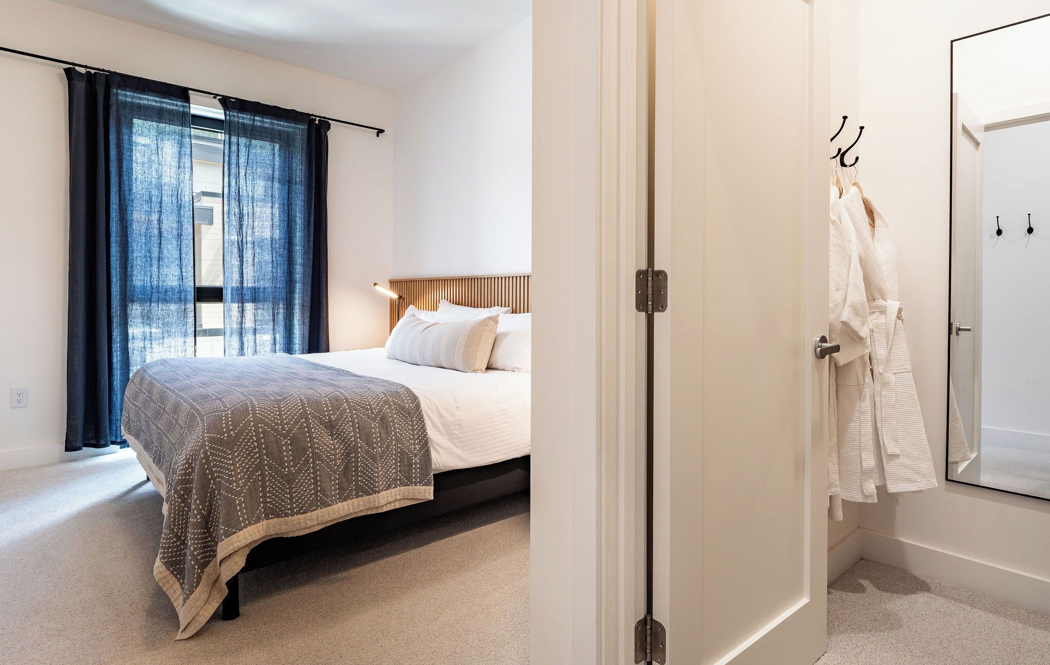 A bedroom with a bed, a bedside table, a window with curtains, and a door leading to a bathroom with a mirror and towel rack.