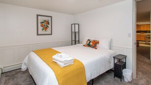 A neatly made bed with a white and yellow blanket, a small bedside table, a black nightstand, a framed picture on the wall, and a view into the adjacent kitchen area. - 4BR I Gameroom, Queen x4, Kitchens x2, Pac n' play (Fargo)