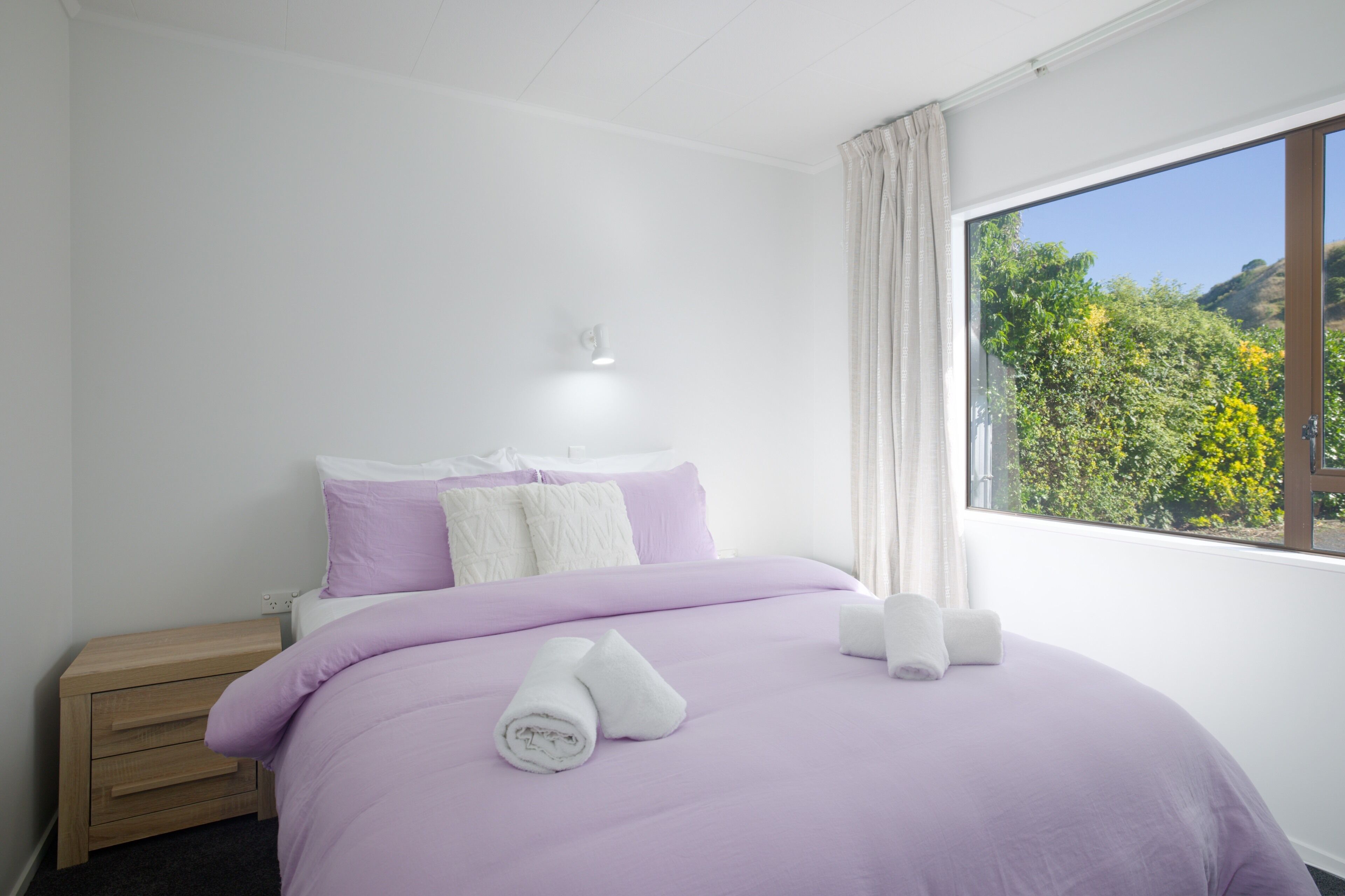A bedroom with a bed, a nightstand, a window with a view of greenery, and a wall-mounted light.