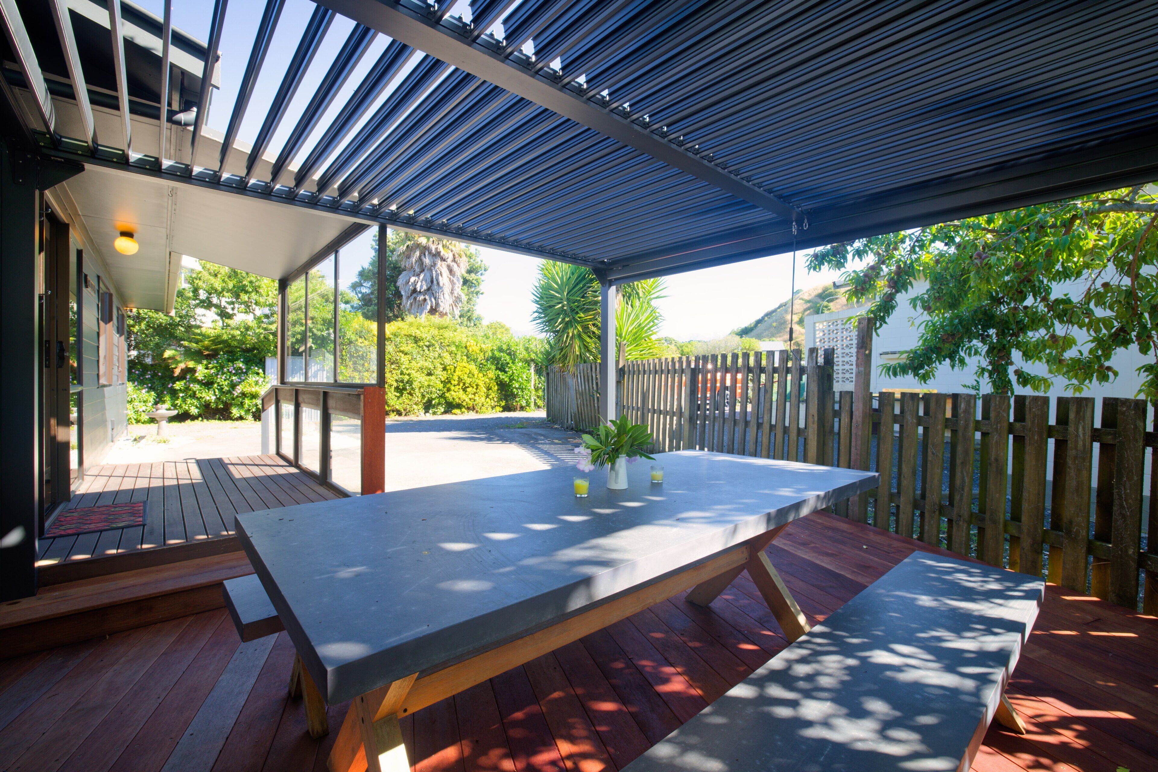 An outdoor patio with a large table and benches, surrounded by a wooden fence and a view of greenery and hills.