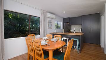 A dining area with a wooden table and chairs, a kitchen area with grey cabinets, and a window with a view of trees.