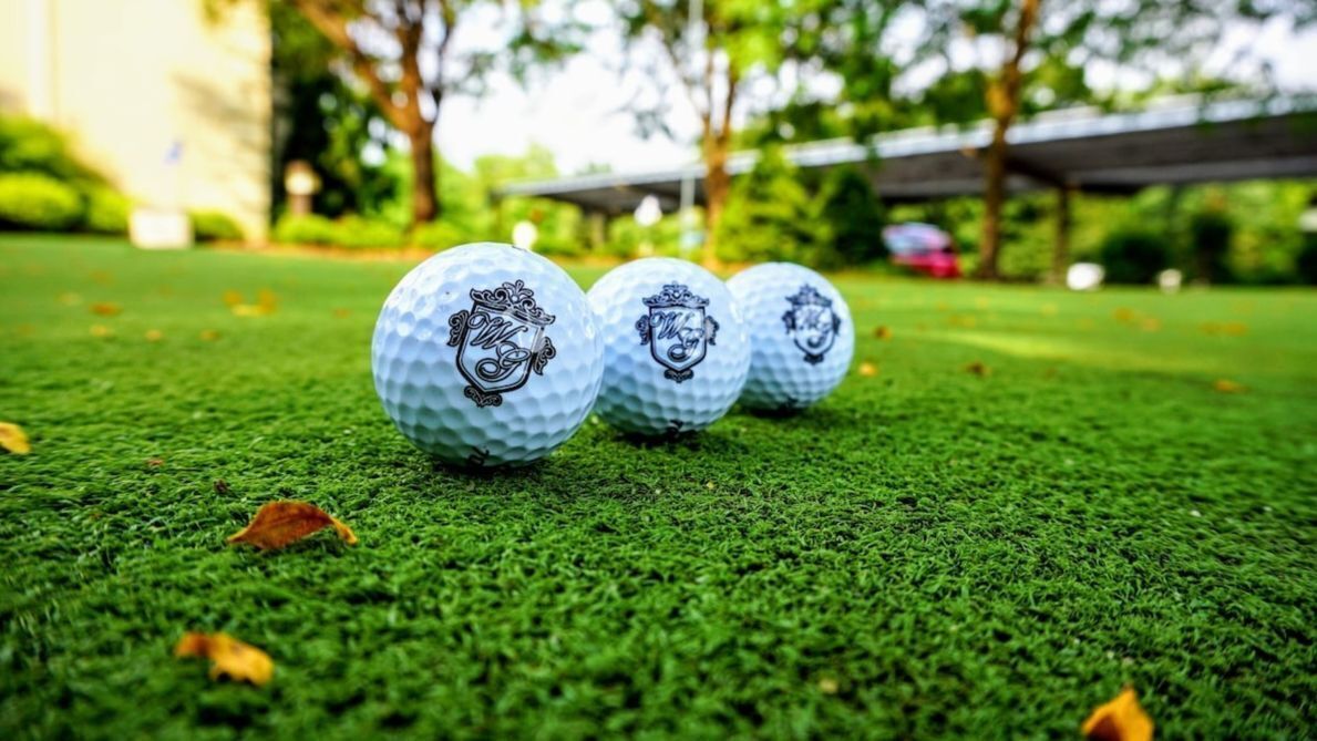 Drei Golfbälle auf einer grünen Fläche, im Hintergrund verschwommen Bäume und ein Gebäude.