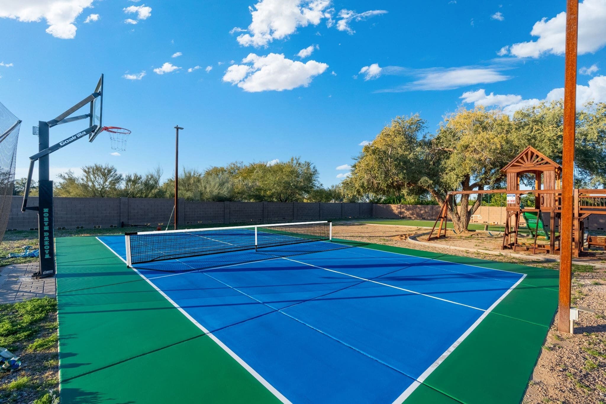 Una cancha de tenis azul y verde con una red, un aro de baloncesto y una estructura de juegos de madera.