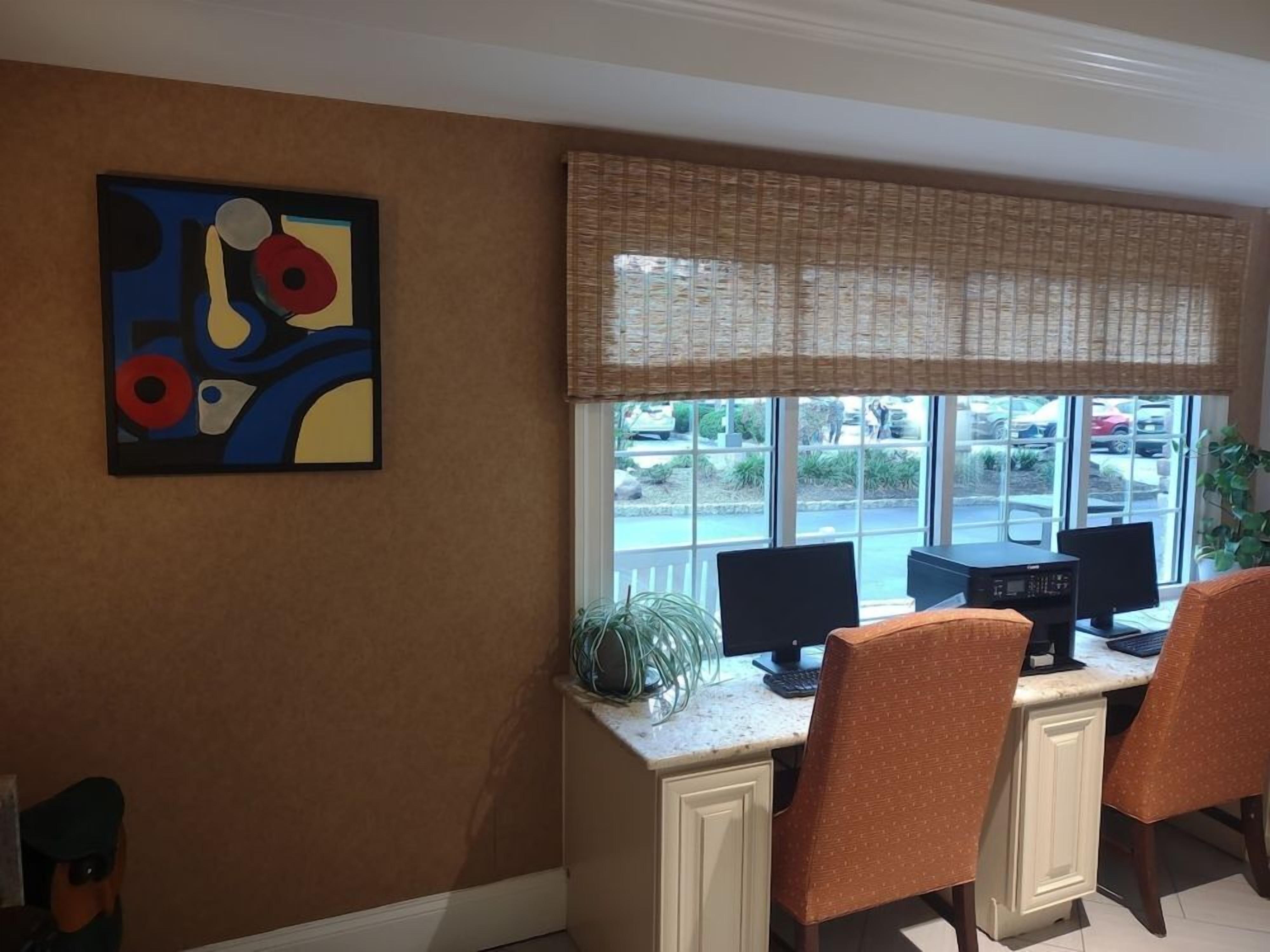 A room with a desk, two monitors, and two chairs. There is a window with a view of a parking lot and a building outside.