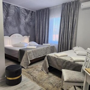 A hotel room with two beds, a small round ottoman, a white chair, and a large window with curtains. - Hotel Mantra (Arad)