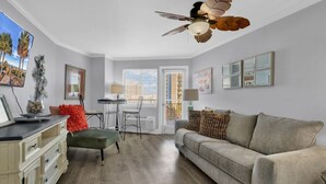 A living room with a beige sofa, a green armchair, a dining table with chairs, a ceiling fan, and a window with blinds. - Bluewater Resort 1BR Condo Steps From Beach (Myrtle Beach)