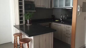 Una cocina con encimera negra, armarios de madera, un microondas, un lavabo y dos taburetes.